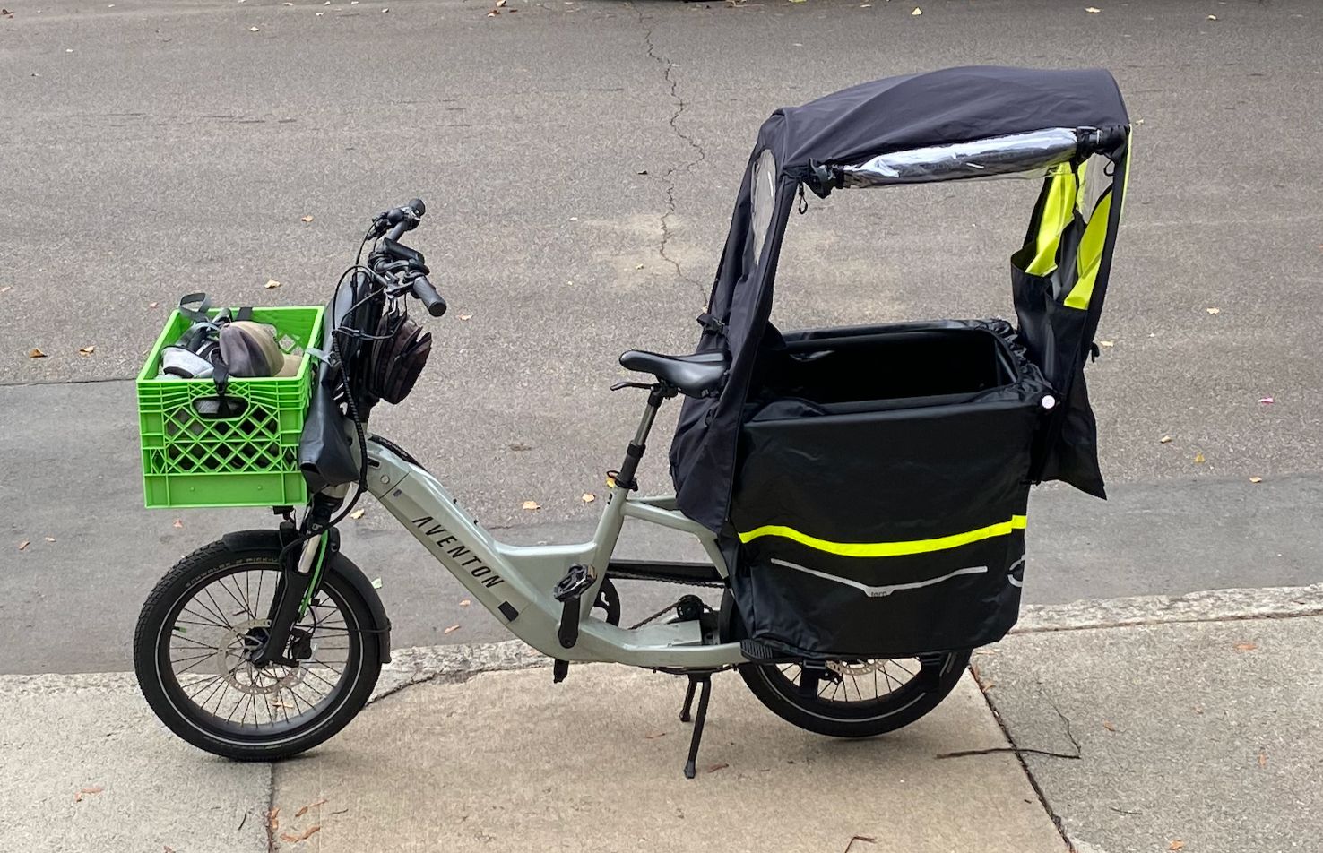 a modern cargo bicycle parked on an asphalt surface. It's equipped with a large black cargo box on the rear, featuring reflective yellow stripes for visibility. The box has a black rain cover with a rolled-up transparent section. Attached to the handlebar is a bright green plastic crate holding various items. The bike is designed for utility and appears ready for all-weather conditions.